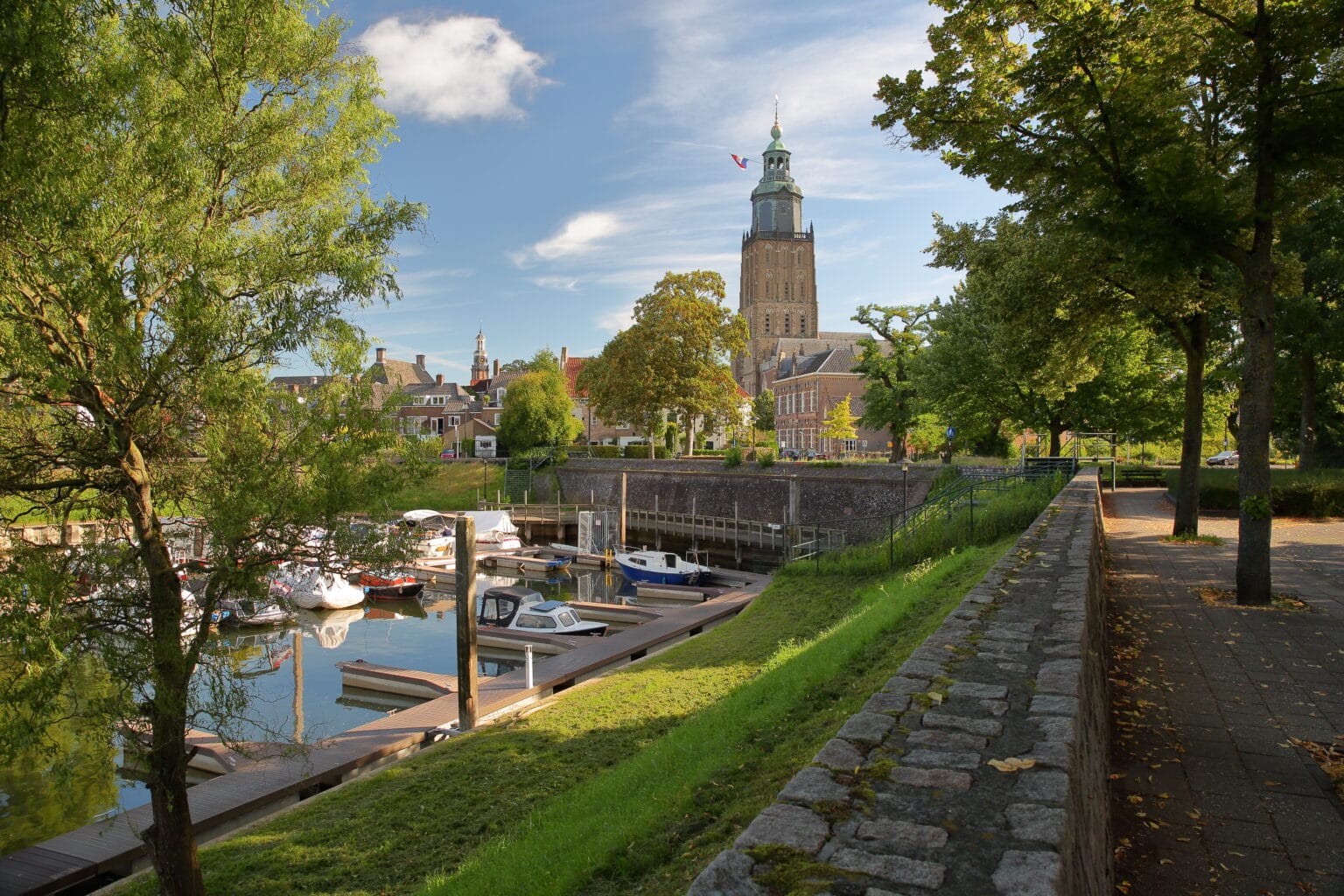 Zonnig uitzicht op de historische haven van Zutphen met aangemeerde boten en de imposante Walburgiskerk op de achtergrond, omringd door groene bomen en karakteristieke oude gebouwen – historic harbor, boats, and St. Walburgis church in Zutphen, the Netherlands